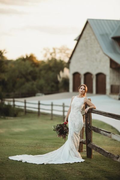 Bride in a lace gown leaning on a wooden fence outside a stone barn at sunset